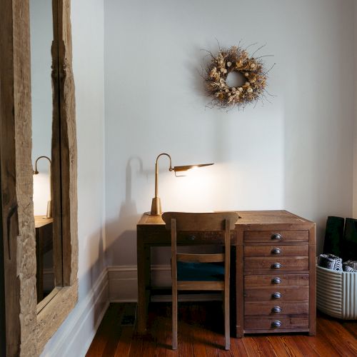 A cozy nook with a wooden desk, chair, and a lamp. A wreath decorates the wall, and a large mirror reflects the space.