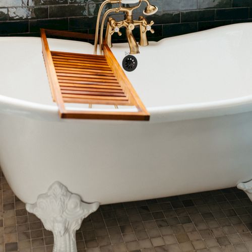 A white clawfoot bathtub with a wooden tray and elegant brass fixtures against dark green tiled walls.