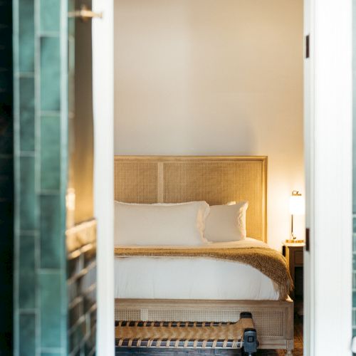 The image shows a cozy bedroom with a woven headboard, neatly arranged bed, soft lighting, and a decorative rug on the floor.