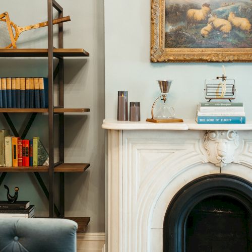 A cozy living room with a bookshelf, books, a blue sofa, a white fireplace, and a framed painting above.