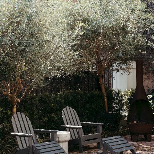 Three Adirondack chairs and a small table on a cobblestone patio, surrounded by trees and greenery in an urban setting.