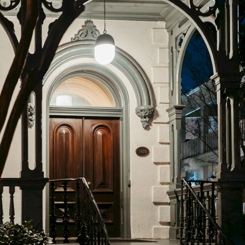 The image shows an illuminated entrance with ornate wood doors and intricate trim, framed by an arch and steps leading up to it.