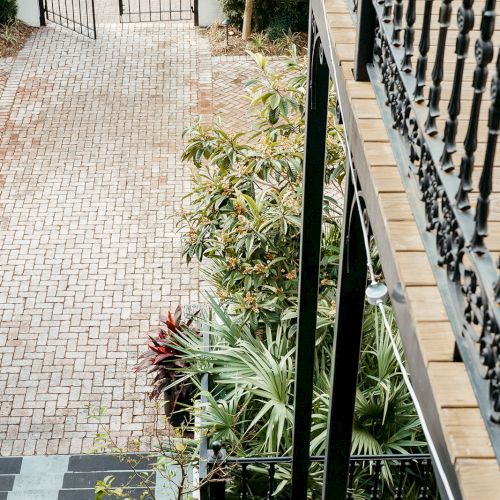 A brick pathway leads to a gate, bordered by potted plants and greenery. A metal railing lines the upper right of the image.