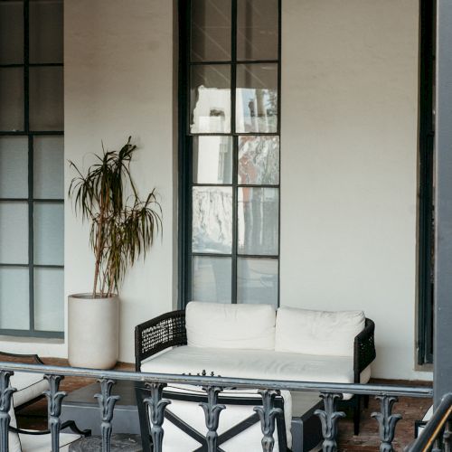 A cozy outdoor patio with a white sofa, a potted plant, and two tall windows, surrounded by a black railing.