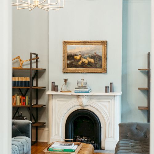 A cozy living room with a fireplace, bookshelves, a chandelier, a painting, and a tufted ottoman in the center of two sofas.