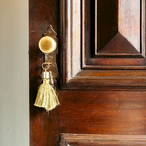 A wooden door is slightly ajar, with a decorated tassel hanging from the doorknob, featuring a small brass bell and a yellow fringe.