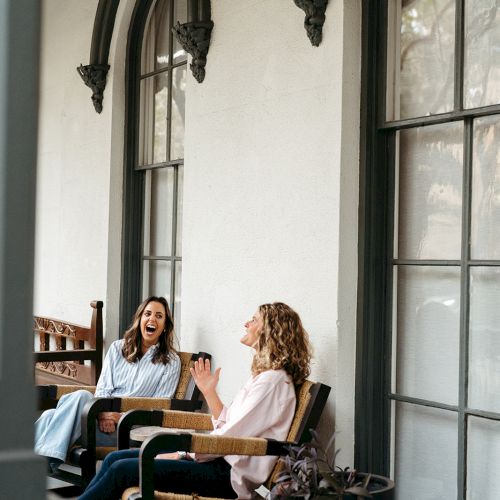 Two people are sitting on a porch, talking and smiling near large windows with arch decorations and a potted plant nearby.