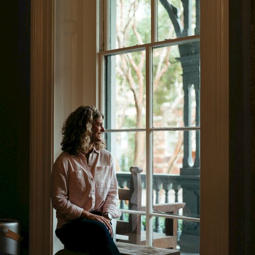 A person sits on a window ledge, looking outside. The setting appears calm and introspective, with a porch and greenery visible beyond.