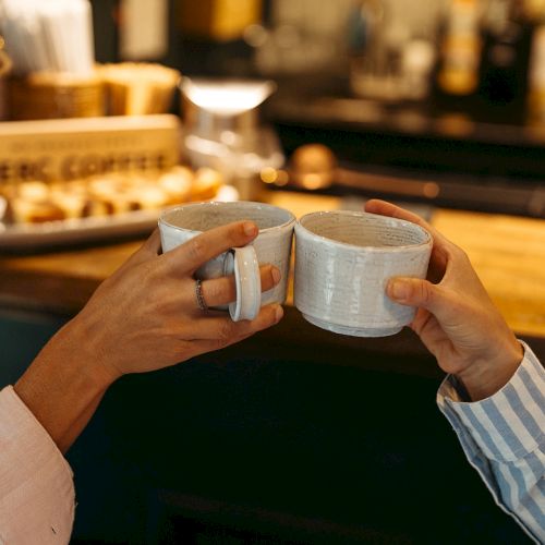 Two people clinking mugs in a cozy coffee shop setting, with a wooden counter and blurred background.