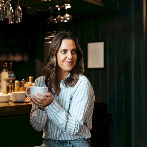 A woman in a striped shirt holds a cup, smiling at another person in a cozy cafe setting.