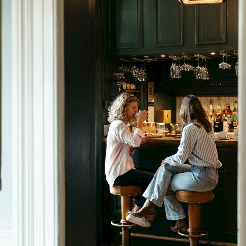 Two people sitting on bar stools, chatting in a dimly lit bar with bottles and glasses visible on shelves behind them.