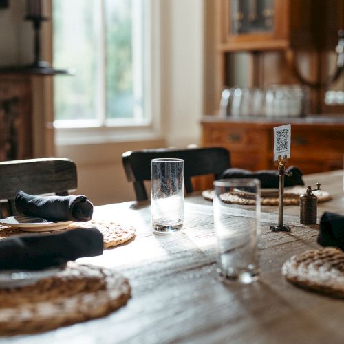 A sunlit dining table set for four with woven placemats, glasses, and napkins, situated in a cozy room with wooden furniture.