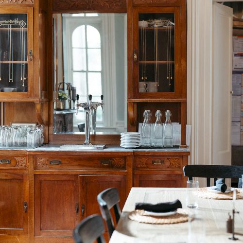 The image shows a dining room with a wooden cabinet and a set table. Glassware and silverware are neatly arranged on both the cabinet and table.