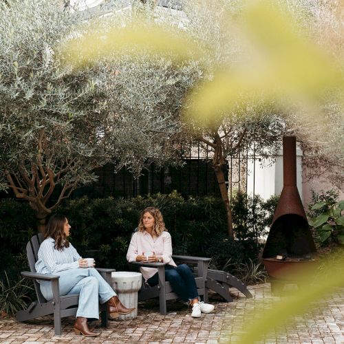 Two people are sitting and talking on outdoor chairs in a garden setting with trees and a fireplace visible.