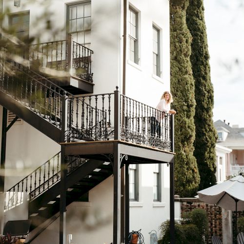 The image shows a white building with a black metal staircase and a balcony. Tall trees are in the background, and a person is on the balcony.