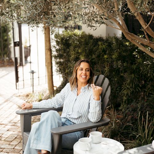 A person is sitting in an outdoor chair, smiling, surrounded by plants and a small table with a drink on it.