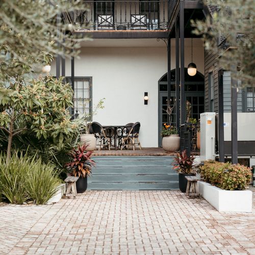 The image shows a building entrance with stairs, plants, and outdoor seating on a brick path.