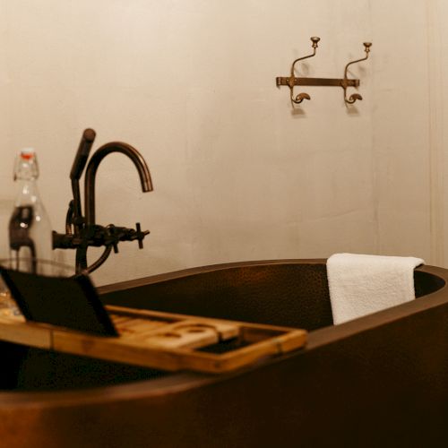 A rustic bathroom with a freestanding tub, bronze faucet, and a wall-mounted towel holder. A wooden bath tray holds items.