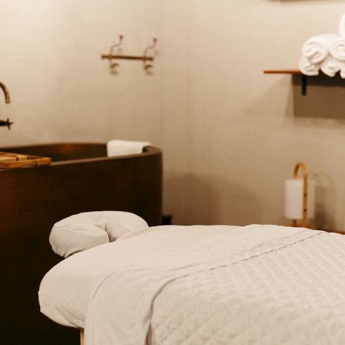 The image shows a spa room with a massage table, rolled towels on a shelf, and a bathtub with a faucet in a serene setting.