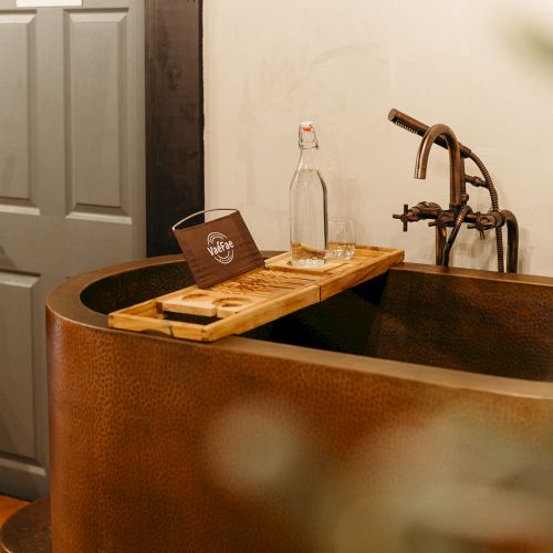 A rustic bathtub with a board holding a bottle and tablet is near a gray door labeled 