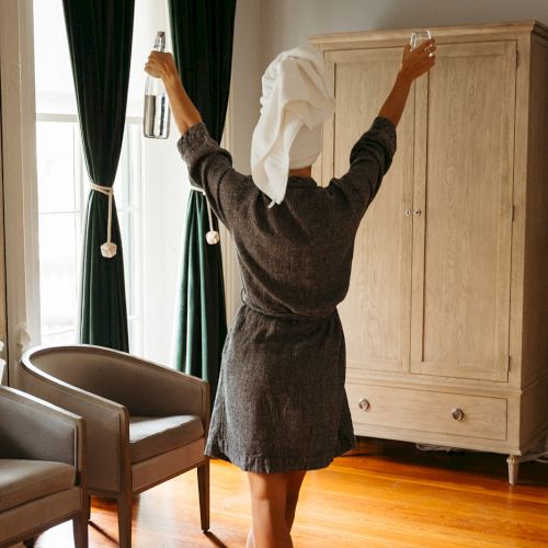 A person with a towel on their head stands in a room, raising their arms, near two chairs and a wooden cabinet.