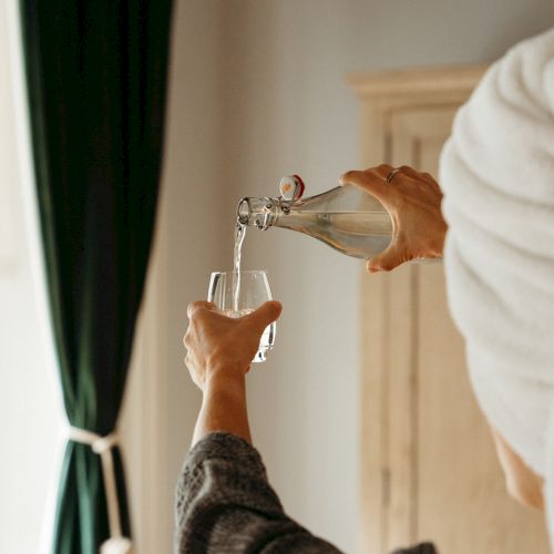 A person with a towel on their head pours clear liquid from a bottle into a glass, with dark green curtains in the background.