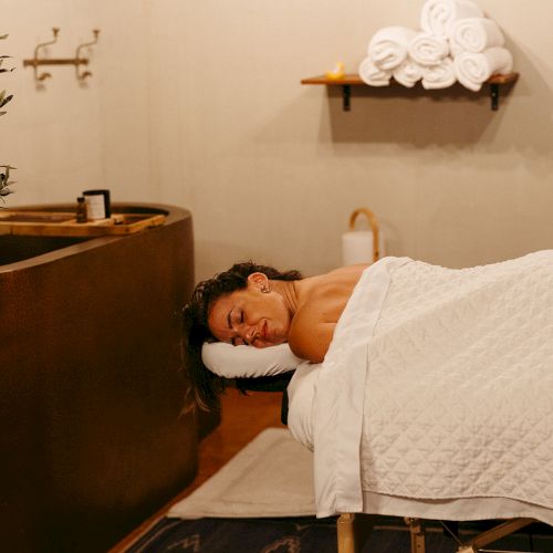 A person is relaxing on a massage table covered with a white blanket. There are rolled towels and a tub in the background.