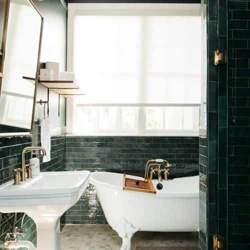 A stylish bathroom with dark green tiles, a white pedestal sink, and a freestanding bathtub with brass fixtures, is shown.