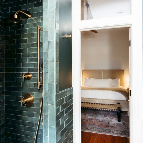 A modern bathroom with green tiles, a gold shower fixture, and a view into a bedroom with a bed and rug through an open door.