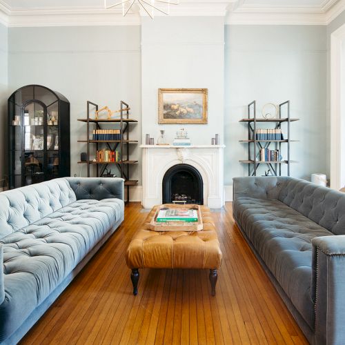 A stylish living room with two blue sofas, a fireplace, bookshelves, and a central ottoman on wooden flooring completes the scene.