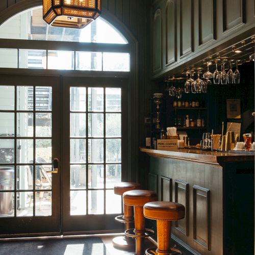 A cozy bar area with wooden stools, a small counter, hanging glasses, and a vintage-style light fixture by a large windowed door.