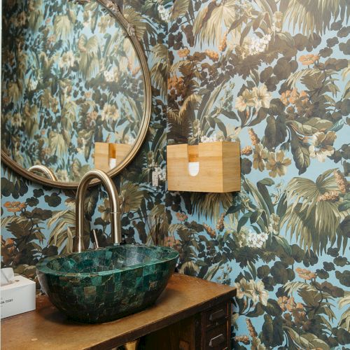 A small bathroom with tropical-themed wallpaper, a round mirror, and a green stone sink on a wooden vanity.