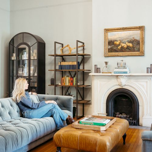 A cozy living room with a woman sitting on a blue sofa, a fireplace, bookshelves, a painting, and a leather ottoman table.