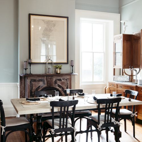 A dining room with a wooden table and six chairs, a chandelier, a fireplace with decor, and a large window letting in light.