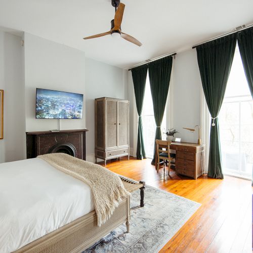 A cozy bedroom with a bed, wooden floor, desk, TV, wardrobe, and large window with green curtains. There's a painting and ceiling fan.