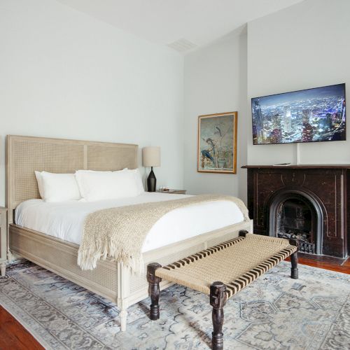 A cozy bedroom with a bed, nightstands, a bench, a TV above a fireplace, and a framed picture on the wall. Elegant and simple decor.