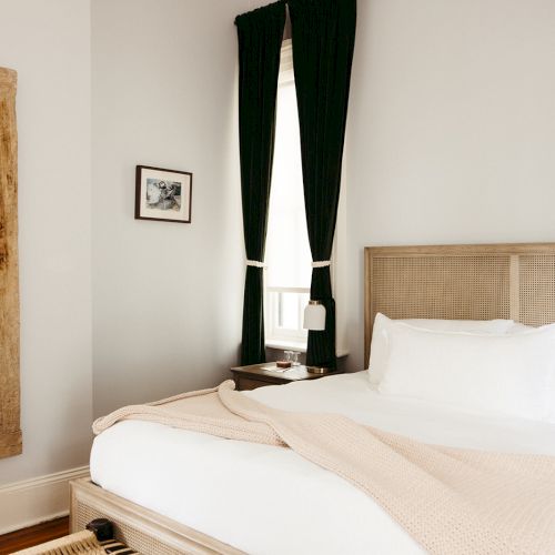 A cozy bedroom with a large bed, green curtains, a wooden-framed mirror, wall art, and a soft blanket draped over the bed.