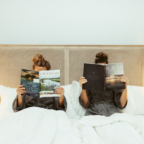 Two people lounging in bed, reading magazines with identical covers, surrounded by cozy lighting and white bedding.