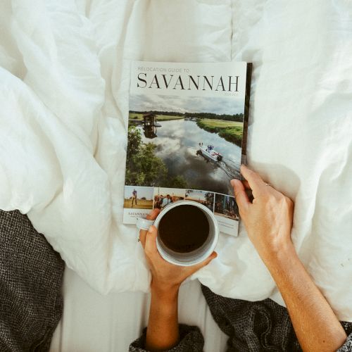 A person holds a magazine titled "Savannah" and a cup of coffee while resting on a bed with white linens.