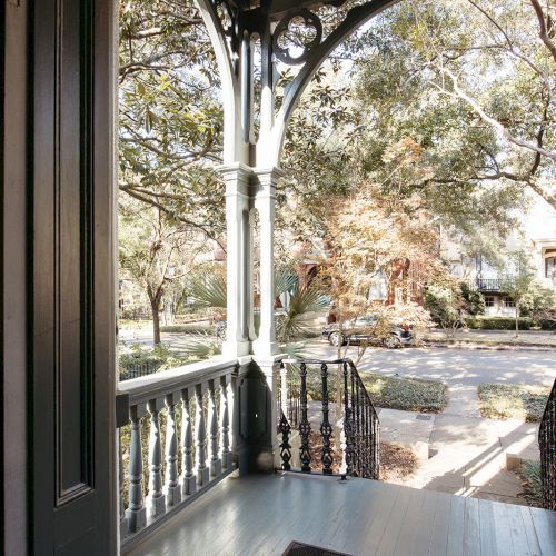 A porch with decorative railings, a hanging light, and a doormat, overlooking a tree-lined street with houses in the background.