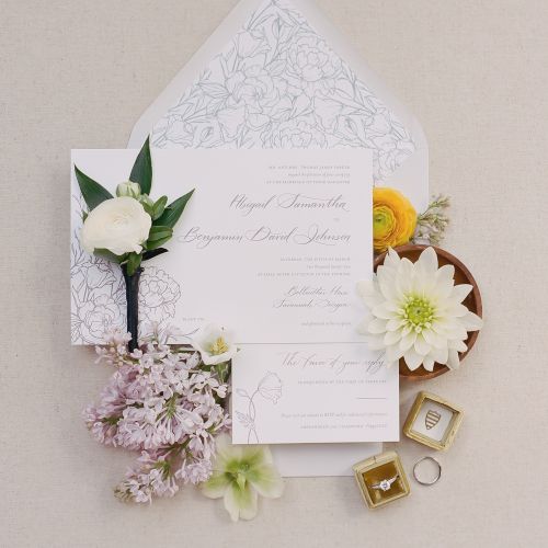 This image features wedding invitations, white flowers, a boutonnière, and two rings in boxes, all arranged on a beige background.