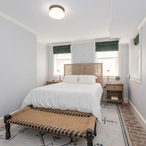 A neat bedroom with a white bed, two nightstands, green window shades, a bench at the foot of the bed, and light walls.