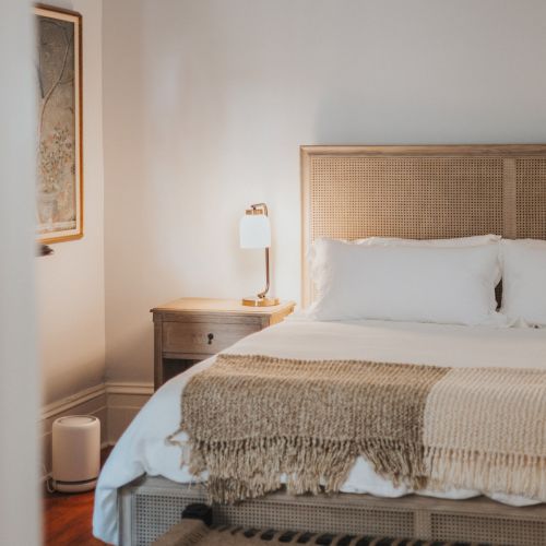 A cozy bedroom with a beige headboard, white bedding, and a knitted throw blanket; a nightstand with a lamp; framed artwork on the wall.