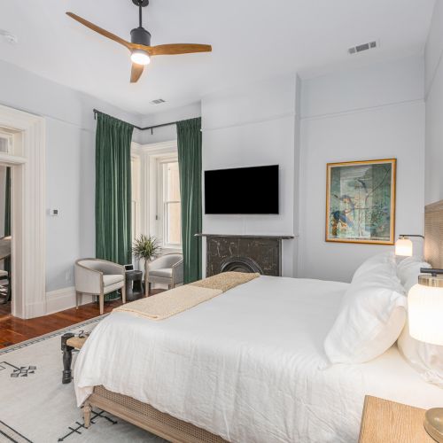 A bedroom with a bed, TV, green curtains, two chairs, a rug, and a fan on the ceiling, with a painting on the wall.