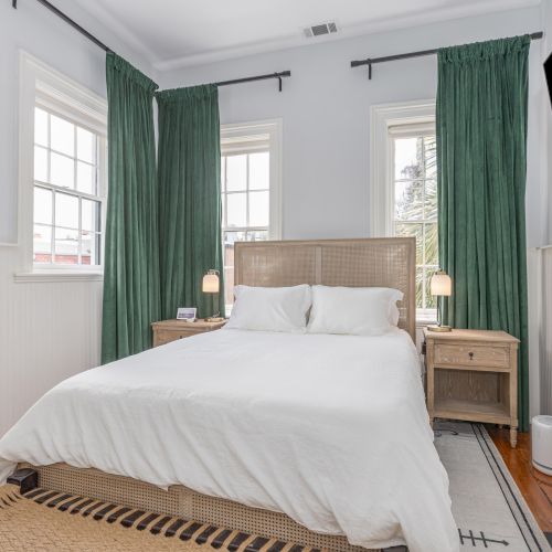 A cozy bedroom features a bed with white linens, green curtains, bedside tables with lamps, a large mirror, and a wall-mounted TV.