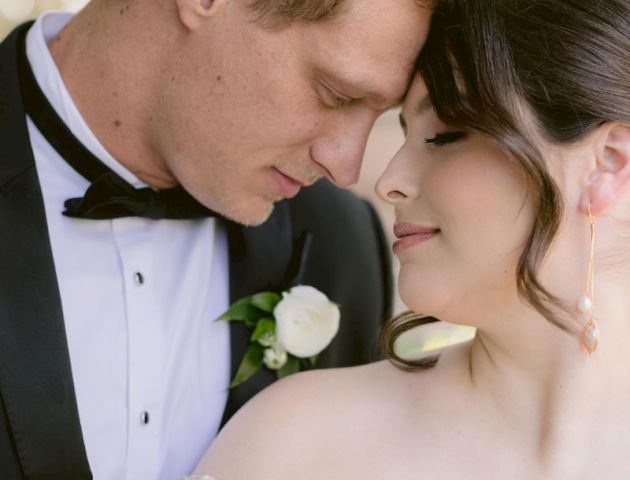 A groom in a black vest kisses a bride on the cheek as she holds a small bouquet in a sunlit outdoor wedding scene.