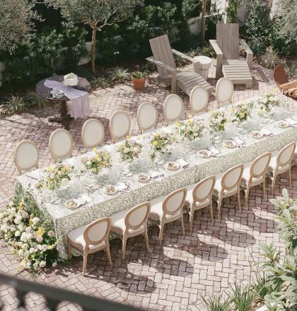 Outdoor garden wedding setup with a long table, floral runner, plates, chairs, side tables, and trees in the background.