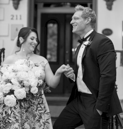 A smiling couple, with the man in a suit and the woman holding a bouquet of flowers, stands in front of a building entrance marked "211."