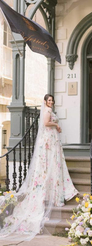 A woman in a floral wedding dress stands on stairs outside a building with black railings, flowers, and a flag that reads "Aristide 211.".