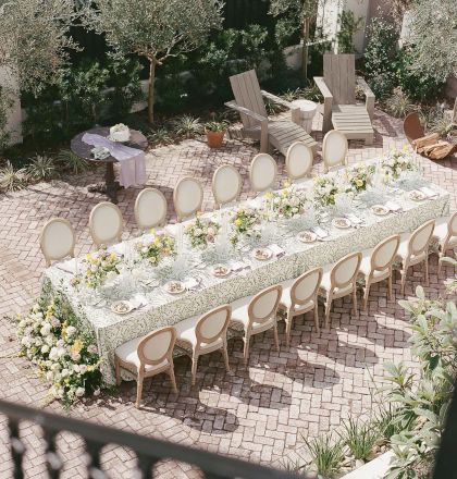 The image shows an elegantly set outdoor dining table with floral arrangements, surrounded by chairs, situated on a brick patio.